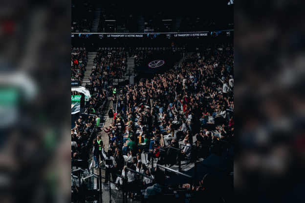 Vue d'ensemble des tribunes de l'Adidas Arena avec les supporters du Paris Basketball célébrant debout dans une ambiance électrique.