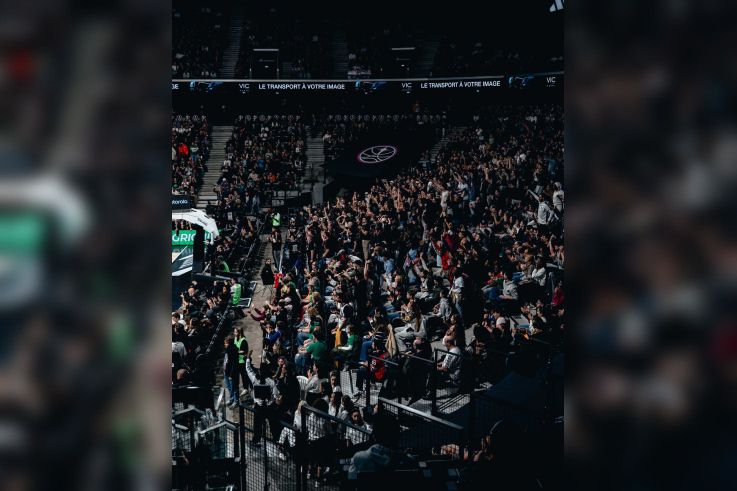 Vue d'ensemble des tribunes de l'Adidas Arena avec les supporters du Paris Basketball célébrant debout dans une ambiance électrique.