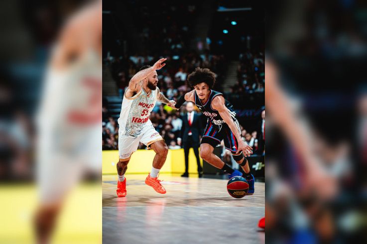 Joueur du Paris Basketball en pleine action offensive, dribblant face à un défenseur lors d'un match de basket intense.